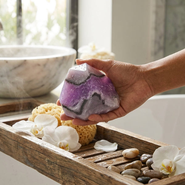 Hand holding a purple and gray amethyst geode soap rock over a wooden tray with bath items in a bathroom setting.